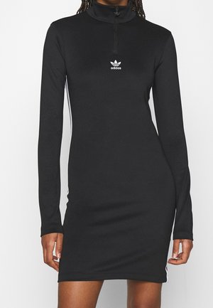 Vrouw draagt een zwarte, aansluitende Adidas-jurk met lange mouwen, een ritssluiting aan de voorkant en een wit logo op de borst.