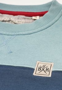 Blå och ljusblå crewneck sweatshirt med ribbad textur, som har en patch med "BXR"-logotyp och röd accent vid kragen.