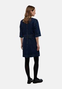 Femme de profil portant une robe en jean foncé avec des manches jusqu'au coude, des collants noirs et des mocassins noirs sur un fond blanc.