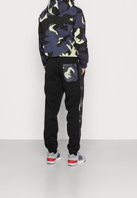 Felpa con cappuccio con motivo mimetico verde e blu navy; pantaloni della tuta neri con tasca mimetica e strisce laterali; scarpe da ginnastica grigie con accenti rossi.