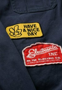 Navy fabric with two embroidered patches: a yellow "Have a Nice Day" patch featuring a peace sign, and a red "Electronic Inc." patch with white text.