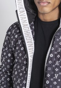 Chaqueta de plumas negra con un patrón de logotipos blancos, cierre de cremallera y una capucha con acentos grises y una tira de marca a lo largo de la cremallera.