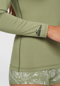 Langarmoberteil in Olivgrün mit Flachnaht und Logo am Ärmel, kombiniert mit gemusterten grünen Shorts mit Blätterdesign.