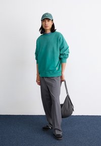 Grøn oversized sweatshirt med ribbede manchetter, grå plisserede bukser, sorte casual sko og en tekstureret grå taske, sat mod en lys baggrund.