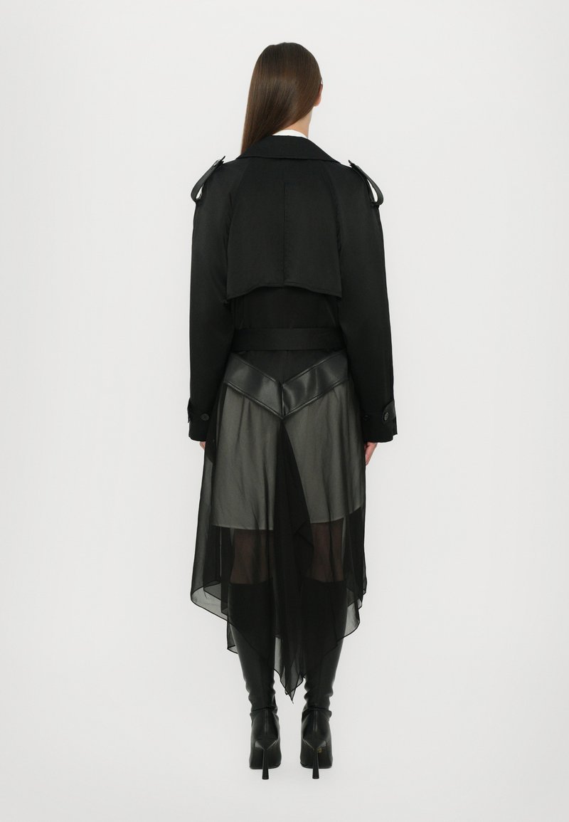 Trench coat nero a strati con parte anteriore corta e schiena lunga e trasparente. Presenta una vita aderente, dettagli lucidi e una silhouette snella.