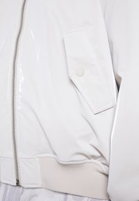 Bílá lesklá bomber bunda se zapínáním na zip, žebrovanými manžetami a našitou kapsou. Hladká textura s jednoduchým designem a jemnými kovovými doplňky.