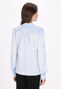 Ljust blå blus med prickigt mönster, genomskinligt material, långa ärmar och krage. Baksidan har veck för extra textur.