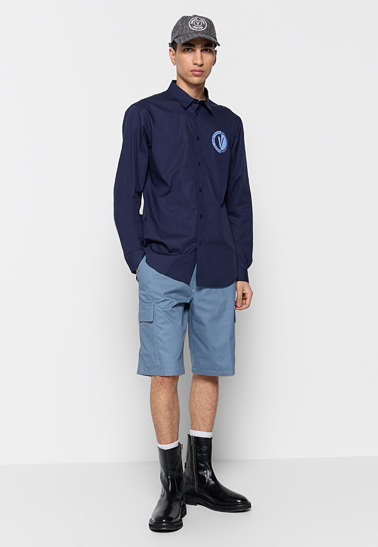 Chemise à manches longues navy avec logo, shorts cargo bleu clair et bottes cheville noires. Une casquette grise avec logo complète la tenue.