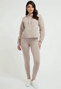 Kvinde står iført en beige Guess hoodie og matchende tætsiddende leggings med hvide sneakers mod en ensfarvet lys baggrund.