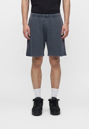 Mand iført mørkegrå knælange shorts, hvide crew-sokker, sorte sneakers og en sort skjorte, stående mod en ensfarvet hvid baggrund.