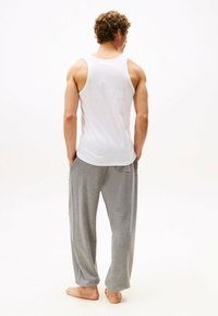 Mann steht barfuß mit dem Rücken zugewandt, trägt ein weißes ärmelloses Shirt und lockere graue Sweatpants mit den Händen in den Taschen.