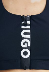 Svart trenings-BH med rund hals, med en sentral glidelås og en iøynefallende hvit "HUGO" logo langs glidelåsbandet. Jevn stofftekstur.