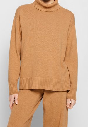 Personne portant un pull à col roulé en maille de couleur chameau avec des manches longues et un pantalon assorti en maille, debout devant un fond gris uni.
