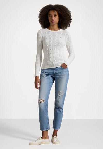 White cable-knit sweater with a round neckline, paired with light blue ripped jeans and beige sneakers. Model stands against a plain background.