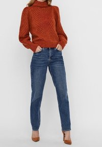 Pull tricoté orange brûlé avec col haut, poignets côtelés et motif texturé, accompagné d'un jean bleu taille haute et de talons marron.