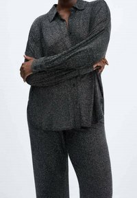 Personne portant un chemisier à manches longues, gris foncé, brillant, avec des boutons et un pantalon assorti, bras croisés sur la poitrine devant un fond uni.