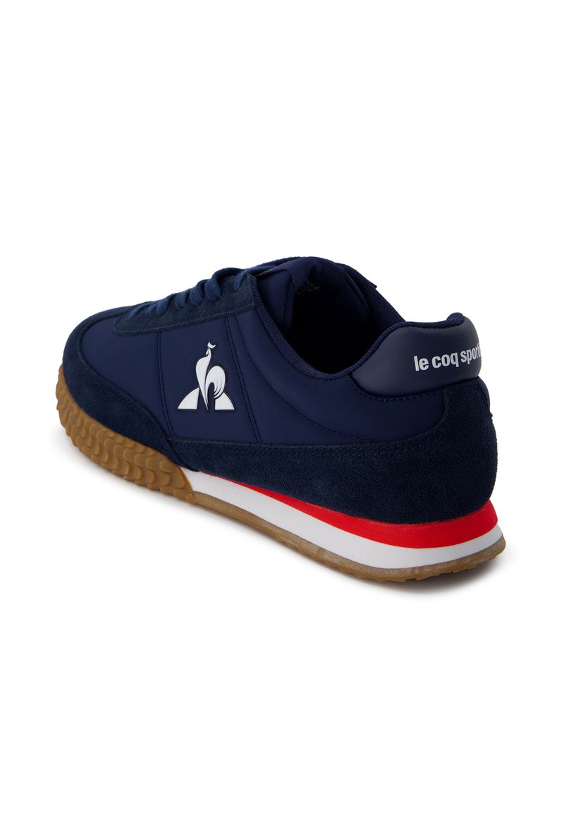 Zapatilla deportiva azul marino con texturas de cuero liso y ante, que presenta un logo blanco, una franja roja y blanca en la suela, y una suela exterior de goma.