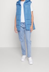 Henkilö, joka wear sleeveless sininen puffer-vesti, valkoinen t-paita, vaaleansiniset farkut ja valkoiset lenkkarit, seisoo yksinkertaista taustaa vasten.