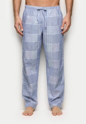 Mann trägt hellblau-weiß karierte Pyjamahosen mit Kordelzug, steht barfuß vor einem schlichten Hintergrund.