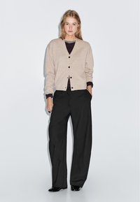 Beige stickad cardigan med v-ringning och svarta knappar, i kombination med vida svarta byxor. Modellen står med händerna i fickorna.