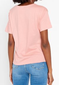 T-shirt a maniche corte, con scollo rotondo, di colore rosa pastello, realizzata in tessuto morbido, mostrata da dietro, abbinata a jeans blu chiaro.