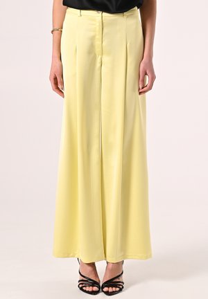 Pantaloni ampi gialli plissè realizzati in tessuto morbido, con vita alta, chiusure frontali e un fit rilassato. Indossati sandali neri con cinturini.