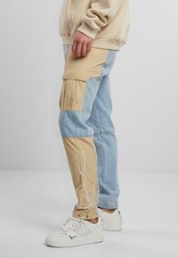 Hellbraune und blassblaue Cargo-Hosen mit Seitentaschen, die ein weißes Liniendesign aufweisen. Kombiniert mit weißen Sneakers, getragen von einer stehenden Person.