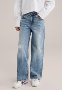 Persona vistiendo jeans de pierna ancha rasgados de color azul claro, camisa blanca de manga larga con puños abullonados y zapatillas blancas con rayas rojas.