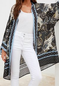Kimono noir et crème à motifs avec des bordures bleu. Présente des motifs floraux et géométriques, en tissu léger et à l'avant ouvert. Porté par-dessus un jean blanc.