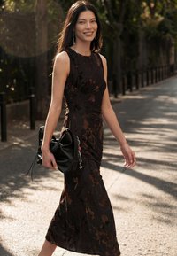 Woman in sleeveless black floral dress walking outdoors holding a black handbag, smiling, with trees and shadows in the background.