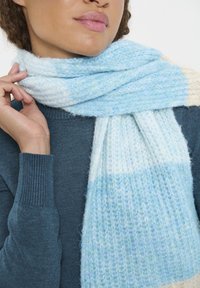 Világoskék sál puha, bolyhos textúrával, különböző kék és fehér árnyalatokkal. Lazaságban viselve a nyak körül, egy sötétkék pulóvert kiegészítve.