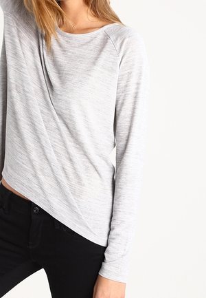 Vrouw draagt een lichtgrijs topje met lange mouwen met een subtiele structuur en zwarte broek, staand tegen een effen witte achtergrond.