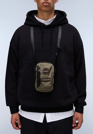 Groene schoudertas met een rits, circulair logo en zwarte accenten, gedragen over een zwarte hoodie met een gestructureerd patroon.