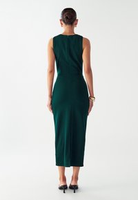 Zelena haljina bez rukava s prilagođenim siluetom, dužine do koljena, i suptilnim detaljem zatvarača na leđima, izrađena od glatkog, teksturiranog materijala.