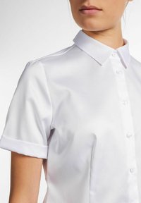 Chemise blanche à manches courtes avec un bouton sur le devant et un col classique. Texture lisse avec des poignets retroussés et un léger éclat. Aucune motif visible.