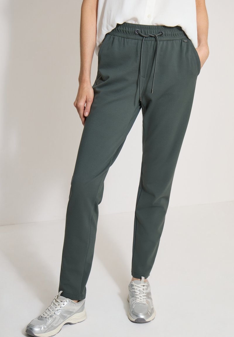 Personne portant un pantalon vert foncé à cordon et des baskets blanches, debout avec une main dans la poche devant un fond uni.