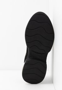 Semelle en caoutchouc noir de chaussure avec un motif de bande de roulement ondulé et le nom de la marque en relief sur la voûte plantaire, positionnée contre un fond blanc d'angle.