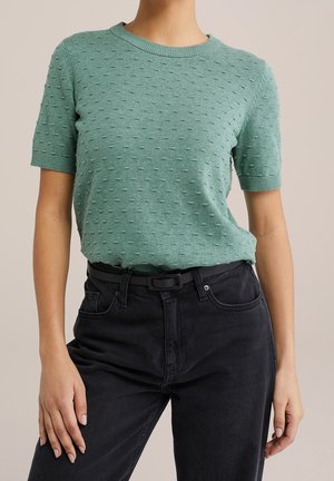 Short-sleeve green knit sweater with textured, raised dots. Round neckline, fitted style, and paired with black high-waisted jeans and a black belt.