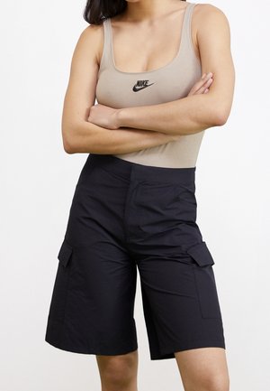 Kobieta w beżowej koszulce na ramiączkach Nike i czarnych szortach bojówkach do kolan, z założonymi skrzyżowanymi rękami, na tle białego, jednolitego tła.