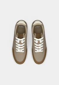 Béžové tenisky s perforovanou ekologickou kůží, hnědými akcenty, bílými tkaničkami a texturovanou podešví. Obsahují polstrovaný límec a logo na jazyku.