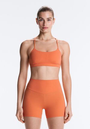 Femme debout face à la caméra, portant un soutien-gorge de sport orange assorti et un short taille haute sur un fond clair uni.