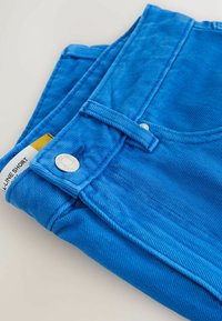 Short en denim bleu vif plié avec fermeture à bouton argenté et étiquette visible portant la mention « A-line Short » sur un fond clair.