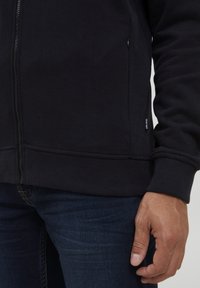 Svart zip-up hettegenser med ribbestrikkede mansjetter og kant, har sidelommer og en liten logoetikett nederst til venstre. Strukturert stoff.