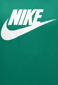 Λευκή επωνυμία Nike και λογότυπο Swoosh τοποθετημένα στο κέντρο σε πράσινο υφασμάτινο φόντο, πιθανότατα μέρος ενός ενδύματος.