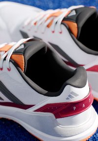 Witte sportieve schoenen met grijze en zwarte accenten, oranje details en een gestructureerde mesh bovenkant. Voorzien van een gevoerde boord en een contrasterende zool.