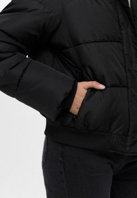 Giubbino invernale nero con texture trapuntata, polsini a coste e vita elastica. Tasca laterale visibile, con chiusura a zip.