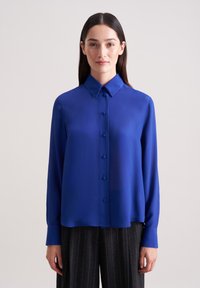 Blaue Seidenbluse mit klassischem Kragen, Knopfverschluss vorne und langen Ärmeln, kombiniert mit grauen Nadelstreifen-Hosen.