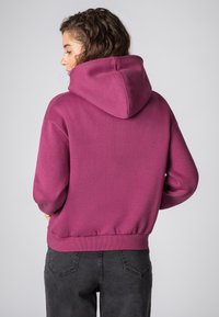 Bordeaux hoodie met een ruime capuchon, geribbelde zoom en manchetten. Zachte textuur en een ontspannen pasvorm, gecombineerd met donkergrijze jeans.