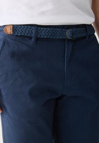 Navy blue cotton trousers with a woven navy belt featuring a brown leather accent. Smooth texture with side pockets and tailored fit details.
