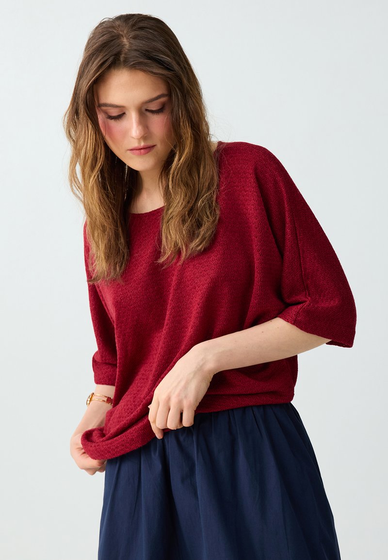 Femme portant un haut rouge texturé à manches courtes ample et une jupe plissée bleu marine, ajustant l'ourlet du haut avec une montre au poignet gauche.